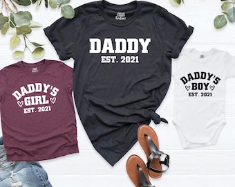 May include: A black t-shirt with white text reading DADDY EST. 2021, a maroon t-shirt with white text reading DADDY'S GIRL EST. 2021, and a white baby bodysuit with black text reading DADDY'S BOY EST. 2021, all featuring heart graphics.