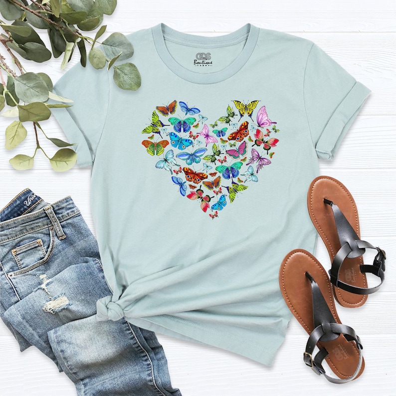 May include: A light blue t-shirt featuring a heart-shaped design made up of colourful butterflies. The butterflies are in various shades of blue, green, yellow, orange, red, and purple.
