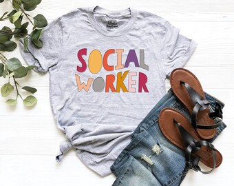 May include: A heather gray short-sleeve t-shirt featuring the text SOCIAL WORKER in multi-colored, wavy-edged block letters. The shirt is paired with blue denim jeans that have distressed details and a pair of brown leather thong sandals.