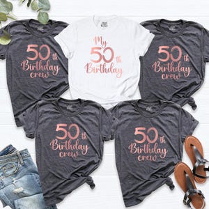 May include: A collection of t-shirts, including a white shirt and several dark gray shirts. The shirts feature the text "My 50th Birthday" or "50th Birthday crew" in a rose gold script. The shirts are casual wear.