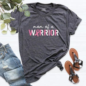 May include: A dark heather gray short-sleeve t-shirt featuring the white text "mom of a" above the word "WARRIOR" in white and pink letters, with a pink breast cancer awareness ribbon replacing the letter "A" in warrior.