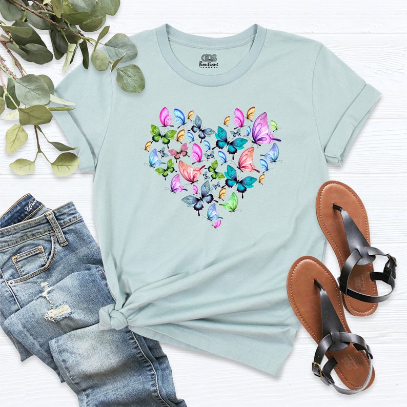 Puede incluir: Una camiseta azul claro con un dise&ntilde;o en forma de coraz&oacute;n formado por mariposas de colores. Las mariposas son de varios tonos de rosa, azul, verde y amarillo.