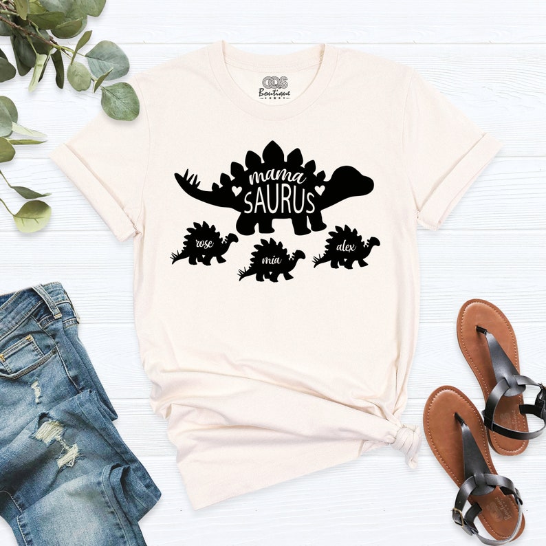 May include: A cream coloured t-shirt with a black silhouette of a dinosaur with the text "mama SAURUS" inside the dinosaur. There are three smaller dinosaurs below the large dinosaur with the names "rose", "mia", and "alex" written below them.