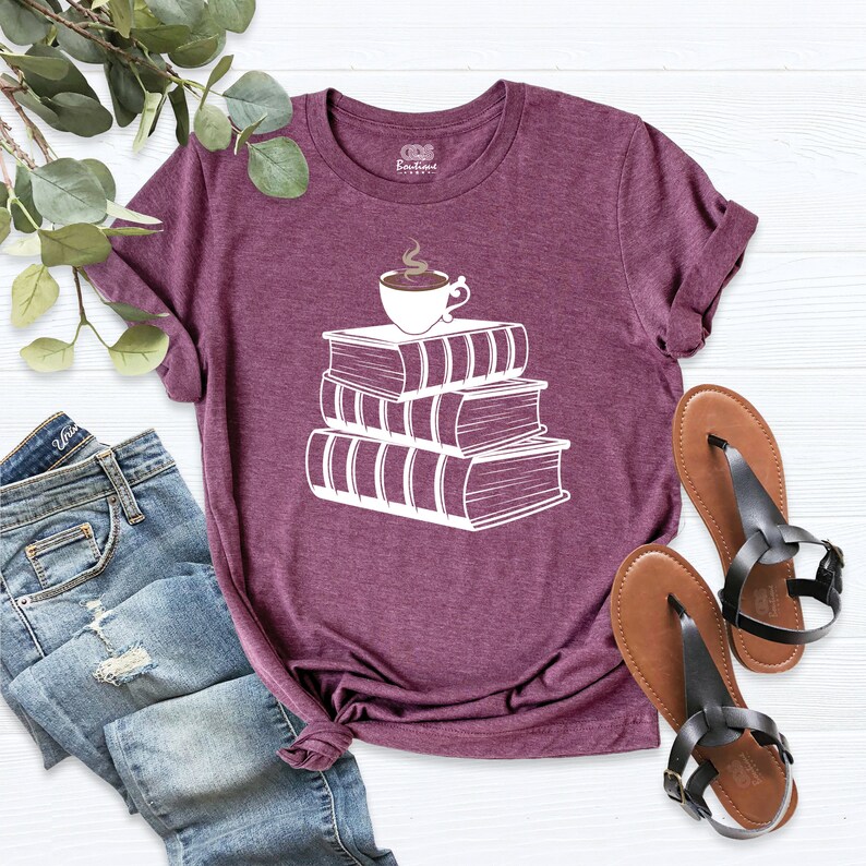 May include: A maroon t-shirt with a white graphic of a stack of books with a steaming cup of coffee on top.
