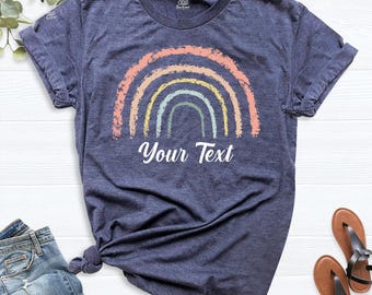 May include: A heather navy blue short-sleeve t-shirt featuring a distressed rainbow graphic in shades of peach, yellow, and sage green. The text "Your Text" is printed in white cursive script centered directly below the rainbow illustration.