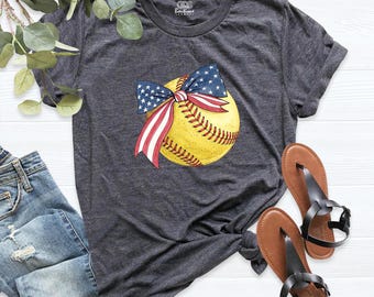 May include: A dark heather gray short-sleeve t-shirt featuring a graphic of a yellow softball adorned with a patriotic bow patterned with red, white, and blue stars and stripes. The shirt has a crew neckline and a knotted hem detail at the side.