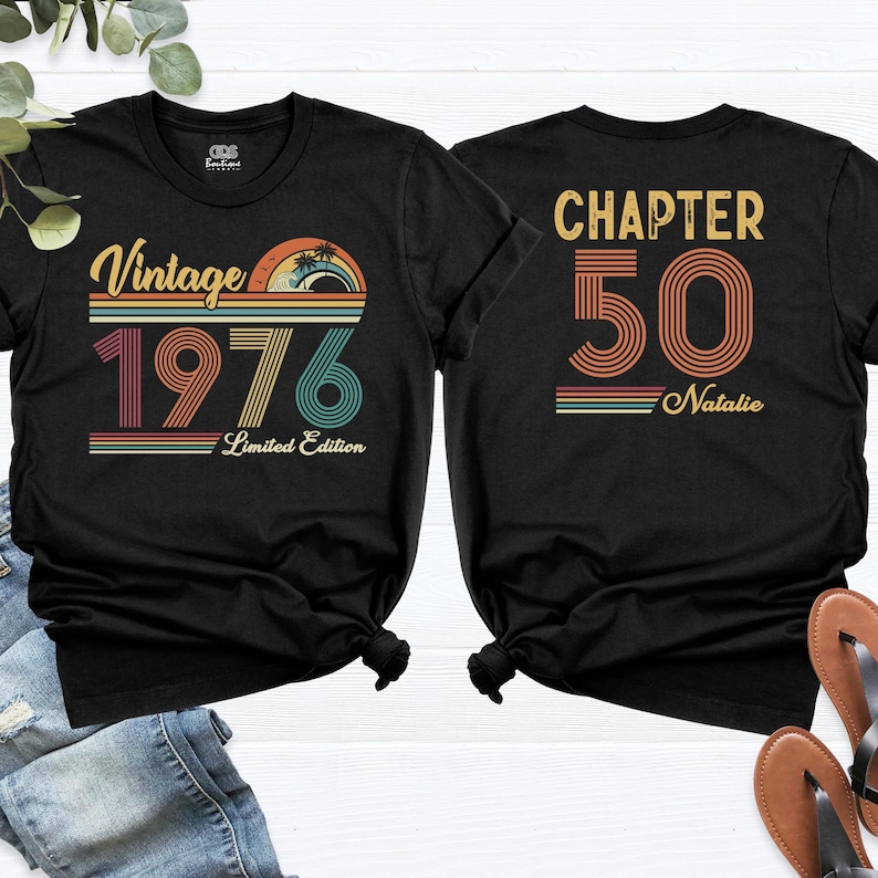 May include: Black t-shirts with retro-style graphics. One shirt features "Vintage 1976" with a sunset design, the other says "Chapter 50 Natalie". Both shirts are short-sleeved and have a crew neck.
