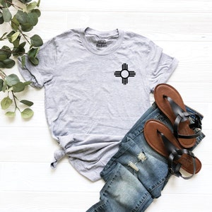 K&ouml;nnte beinhalten: Ein graues T-Shirt mit einem schwarzen Zia-Symbol auf der Vorderseite. Das Shirt liegt auf einer wei&szlig;en Holzoberfl&auml;che mit einem Paar braunen Sandalen und einer blauen Jeans.