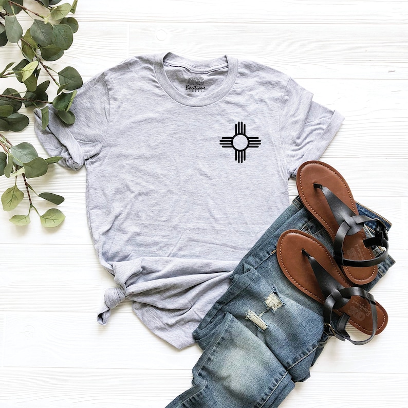 K&ouml;nnte beinhalten: Ein graues T-Shirt mit einem schwarzen Zia-Symbol auf der Vorderseite. Das Shirt liegt auf einer wei&szlig;en Holzoberfl&auml;che mit einem Paar braunen Sandalen und einer blauen Jeans.