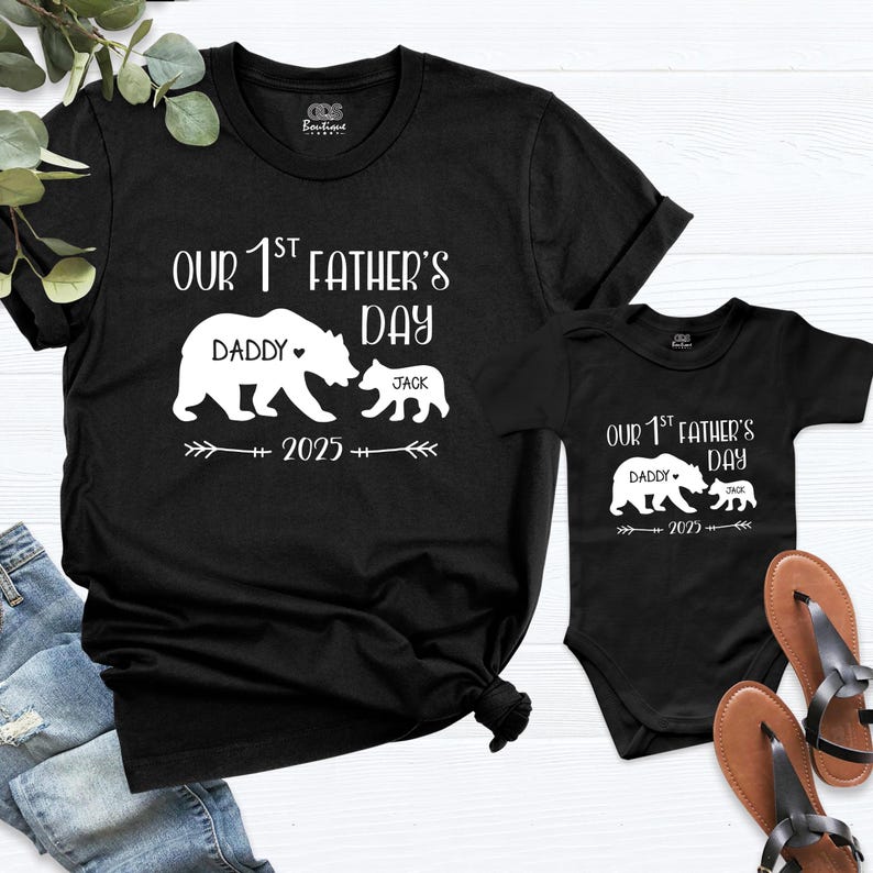 Pu&ograve; includere: Magliette nere e un body con grafica bianca. La grafica recita "Our 1st Father's Day" con illustrazioni di orsi e i nomi "Daddy" e "Jack". &Egrave; incluso anche l'anno "2025".
