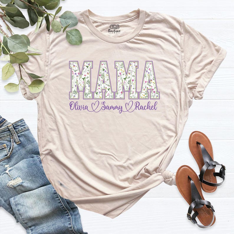 Puede incluir: Una camiseta beige con la palabra "MAMA" en letras grandes de color morado con un patr&oacute;n floral. Los nombres "Olivia", "Sammy" y "Rachel" est&aacute;n escritos debajo de la palabra "MAMA" con corazones entre cada nombre.