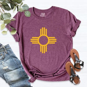 May include: A maroon t-shirt featuring a yellow Zia symbol, a Native American design. The Zia symbol is a circle with four groups of four lines radiating outwards. The shirt is paired with blue jeans and black sandals.