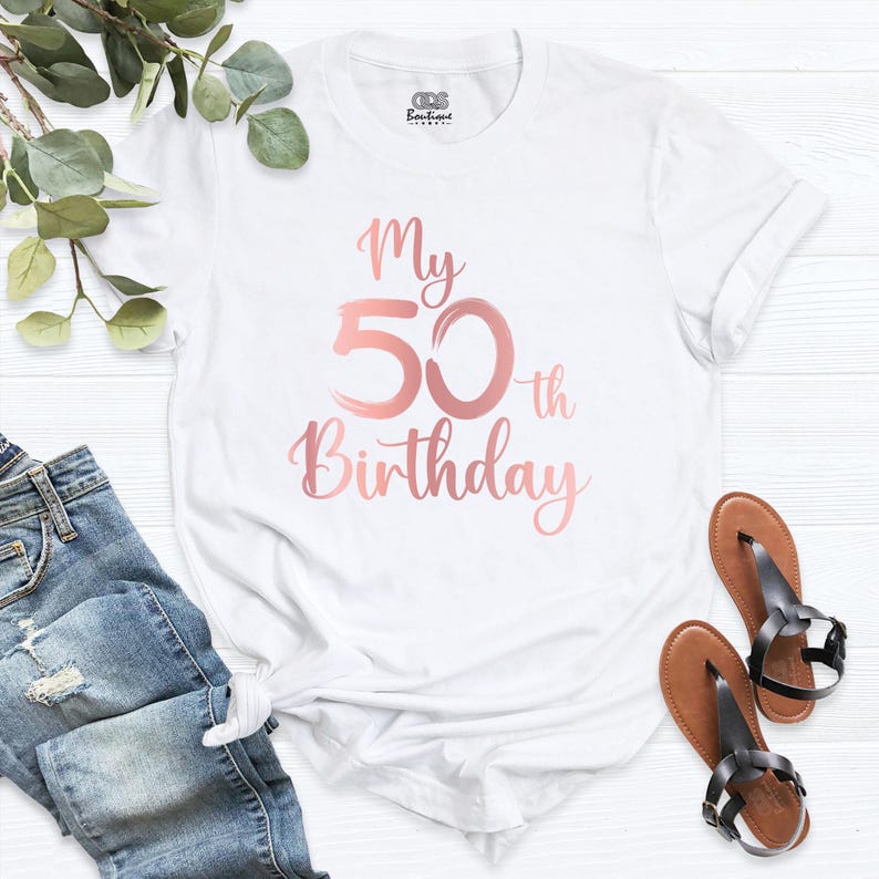 May include: White t-shirt with the text "My 50th Birthday" in rose gold script. The shirt is paired with blue jeans and brown sandals with black straps. A sprig of greenery is in the background.