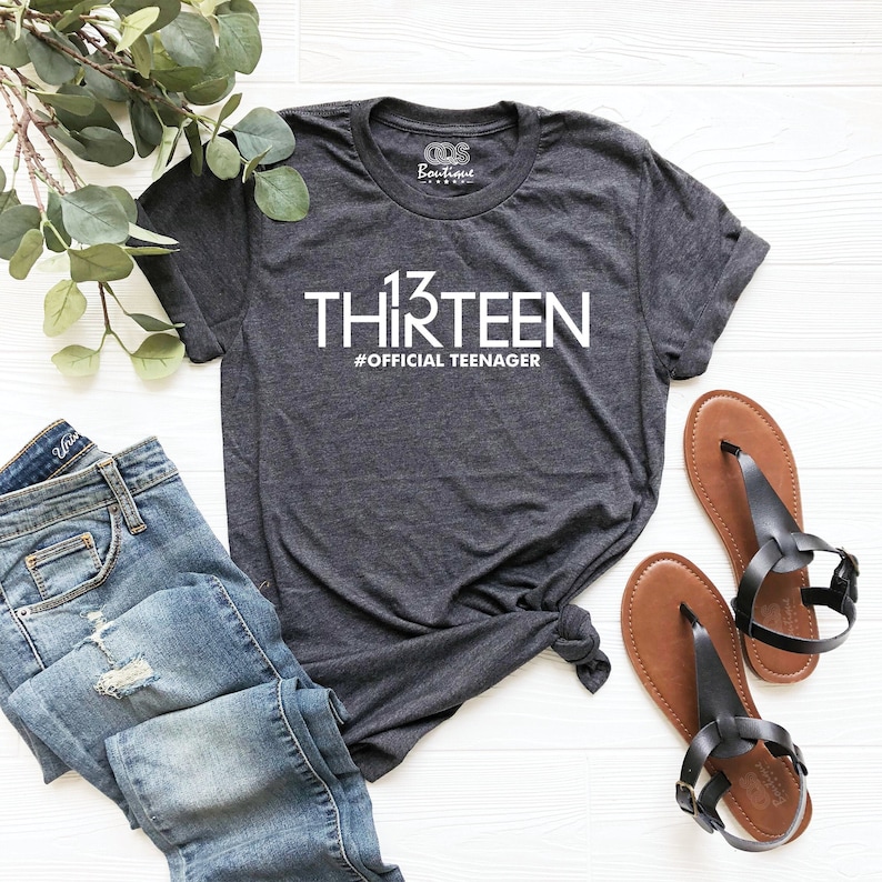 May include: A dark gray t-shirt with white text that reads "13 THIRTEEN #OFFICIAL TEENAGER". The shirt is laid out on a white wooden surface with a pair of brown sandals and blue jeans.