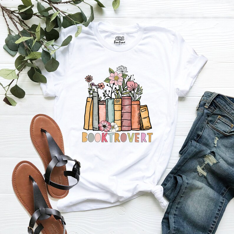 May include: White t-shirt featuring a graphic of a stack of colourful books with flowers on top and the text "BOOKTROVERT" underneath.