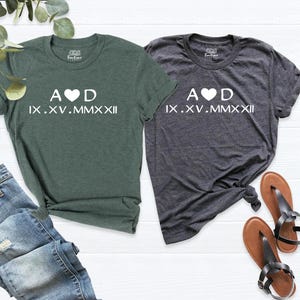May include: Two t-shirts, one green and one grey. The shirts feature a white graphic with the text "A &hearts; D IX.XV.MMXXII".
