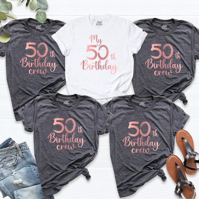 May include: A collection of t-shirts, including a white shirt and several dark gray shirts. The shirts feature the text "My 50th Birthday" or "50th Birthday crew" in a rose gold script. The shirts are casual wear.