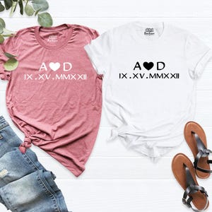 May include: Two t-shirts with a heart and the letters A and D, and the date IX.XV.MMXXII in Roman numerals. The shirts are pink and white.