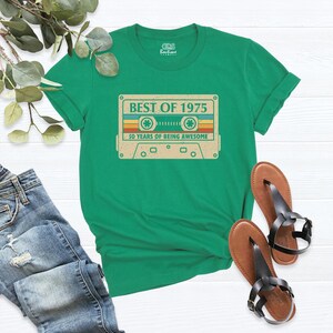 May include: A green t-shirt featuring a retro cassette tape design with the text "BEST OF 1975" and "50 YEARS OF BEING AWESOME". The shirt is paired with blue jeans and brown sandals.