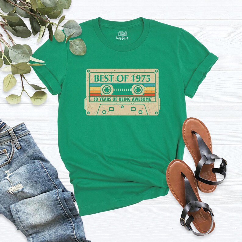 May include: A green t-shirt featuring a retro cassette tape design with the text "BEST OF 1975" and "50 YEARS OF BEING AWESOME". The shirt is paired with blue jeans and brown sandals.