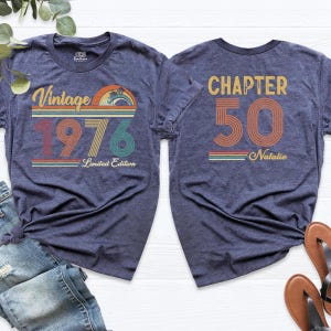 May include: Two navy blue t-shirts. One features a vintage design with "Vintage 1976" and "Limited Edition" text. The other shirt displays "Chapter 50" and "Natalie" text. Both shirts have a retro color scheme.