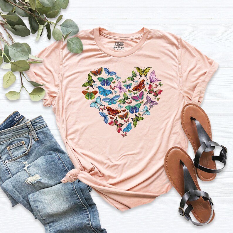 May include: A peach-coloured t-shirt with a heart-shaped design made up of colourful butterflies. The shirt is made of a soft, comfortable material.