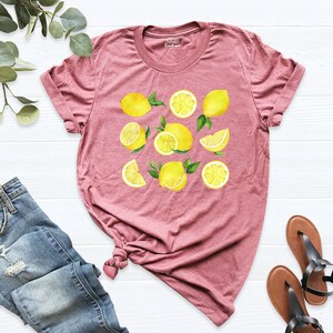 May include: A mauve t-shirt with a lemon print. The lemons are yellow and have green leaves. The shirt is made of a soft, comfortable material.
