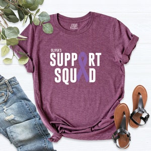 K&ouml;nnte beinhalten: Lilafarbenes T-Shirt mit wei&szlig;em Text, der "Olivia's Support Squad" lautet, mit einem lilafarbenen Band-Motiv.