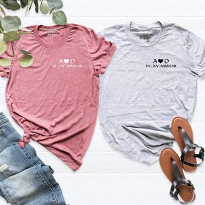 May include: A mauve t-shirt and a heather gray t-shirt, each featuring the text A heart D and the Roman numeral date IX.XV.MMXXII printed on the chest. The mauve shirt has white text, while the gray shirt has black text.