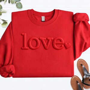 May include: A red sweatshirt with the word "love" embossed across the front. The word "love" is in a bold, sans-serif font, with a small heart replacing the "o". The sleeves are cuffed. Sandals are visible in the bottom right corner.