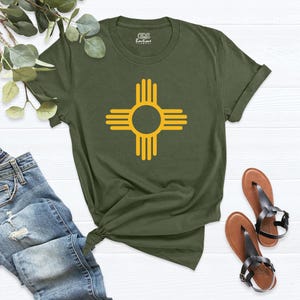 May include: Olive green t-shirt featuring a yellow Zia symbol, a Native American symbol from New Mexico. The shirt has a crew neck and short sleeves. The image also shows a pair of brown sandals and a pair of blue jeans.