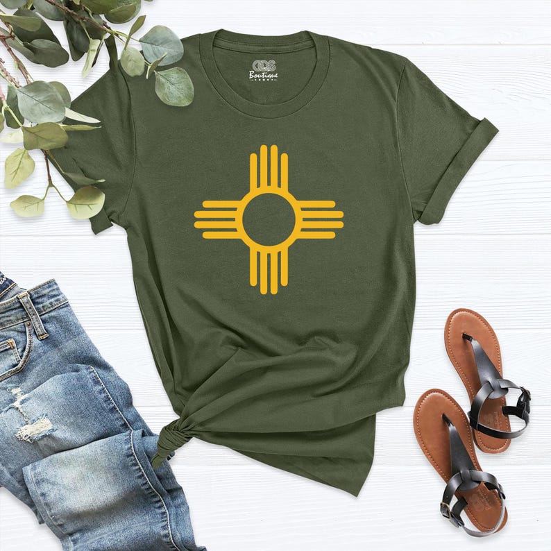 May include: Olive green t-shirt featuring a yellow Zia symbol, a Native American symbol from New Mexico. The shirt has a crew neck and short sleeves. The image also shows a pair of brown sandals and a pair of blue jeans.