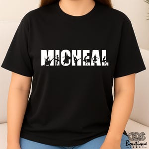 May include: Black t-shirt with the word "MICHEAL" in large white capital letters. The word "MICHEAL" has silhouettes of ballet dancers in various poses. The shirt is made of a soft material and is suitable for casual wear.
