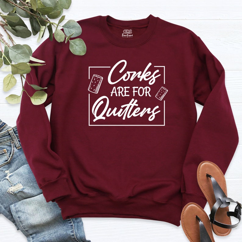 Op de afbeelding: Bordeauxkleurig sweatshirt met witte tekst die "Corks are for Quitters" luidt met een afbeelding van twee wijnkurken.