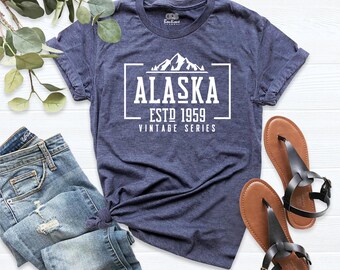 May include: A heathered navy blue short-sleeve t-shirt featuring a white graphic print of a mountain range above the text ALASKA ESTD 1959 VINTAGE SERIES. The shirt is displayed alongside blue denim jeans and a pair of brown leather sandals.