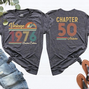 May include: Two charcoal gray t-shirts with retro designs. The front shirt reads "Vintage 1976 Limited Edition" with a colorful sunset graphic. The back shirt says "Chapter 50 Natalie" in a matching retro style.
