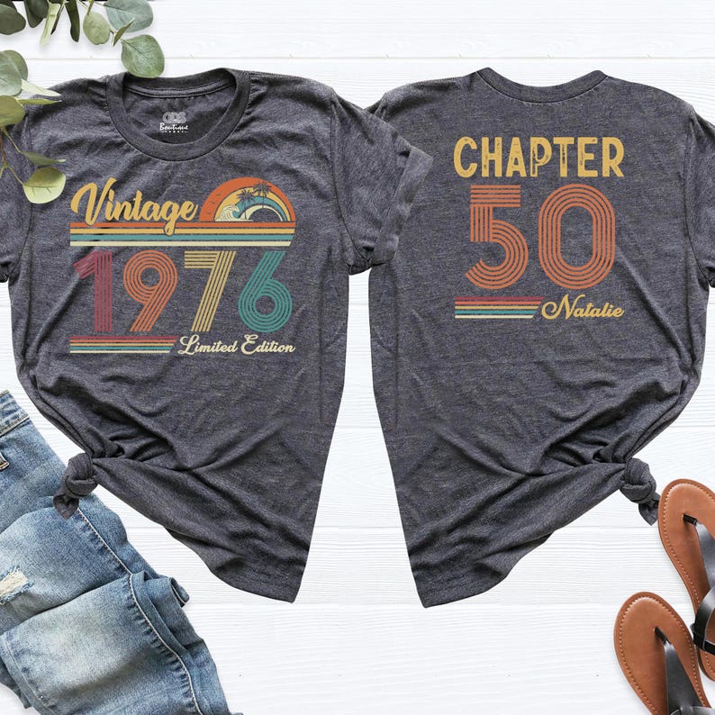 May include: Two charcoal gray t-shirts with retro designs. The front shirt reads "Vintage 1976 Limited Edition" with a colorful sunset graphic. The back shirt says "Chapter 50 Natalie" in a matching retro style.