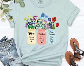 Può includere: Una t-shirt azzurro chiaro con una grafica di tre barattoli di vetro riempiti di fiori di campo colorati. I barattoli riportano le scritte Mom, Grandma e Great-Grandma, ciascuno con un elenco di nomi e un piccolo cuore nero.