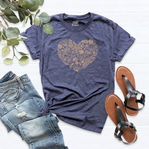 May include: A navy blue t-shirt with a heart-shaped design made of seashells in a light brown color. The t-shirt is laid flat on a white wooden surface with a pair of brown sandals and a pair of blue jeans.