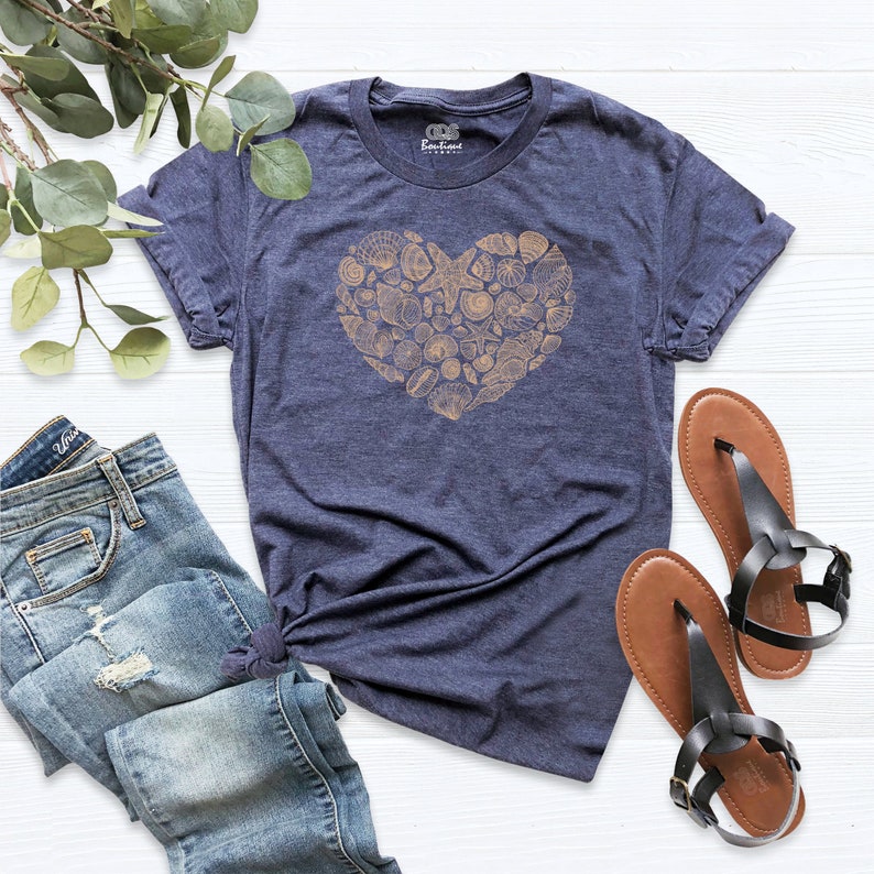 May include: A navy blue t-shirt with a heart-shaped design made of seashells in a light brown color. The t-shirt is laid flat on a white wooden surface with a pair of brown sandals and a pair of blue jeans.