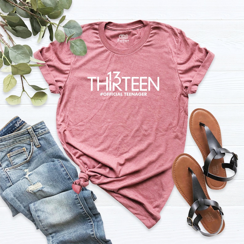 May include: A heather pink t-shirt with white text that reads "13 THIRTEEN #OFFICIAL TEENAGER". The shirt is laid flat on a white surface with a pair of brown sandals and blue jeans.