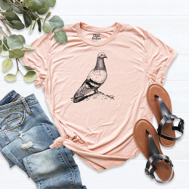Op de afbeelding: Een perzikkleurig T-shirt met een zwart-witte illustratie van een duif op de voorkant. Het T-shirt heeft een ronde hals en korte mouwen.