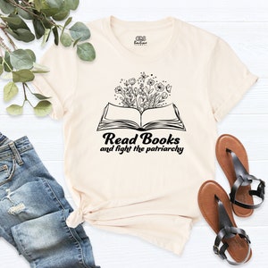 May include: A cream colored t-shirt with a black graphic of an open book with flowers growing out of it. The text "Read Books and fight the patriarchy" is below the book.