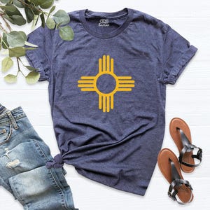 May include: A navy blue t-shirt featuring a yellow Zia symbol, a Native American sun symbol, centered on the front. The shirt is paired with blue jeans and brown sandals.