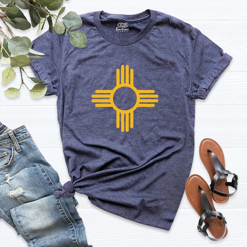 May include: A navy blue t-shirt featuring a yellow Zia symbol, a Native American sun symbol, centered on the front. The shirt is paired with blue jeans and brown sandals.