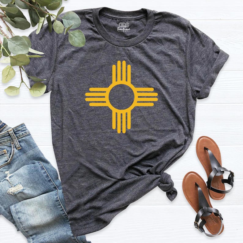 May include: Dark gray t-shirt featuring a yellow Zia symbol, a Native American symbol from New Mexico. The shirt is made of soft material and has a crew neck. The Zia symbol is centered on the front of the shirt. The shirt is paired with blue jeans and sandals.