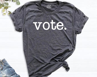 May include: A dark heather gray short-sleeve t-shirt with a crew neckline and rolled sleeves. The word vote. is printed in white lowercase letters across the center of the chest. The shirt features a knotted detail on the lower right side.