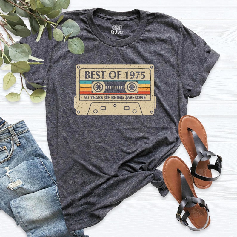 May include: Dark gray t-shirt featuring a retro cassette tape design with the text "BEST OF 1975" and "50 YEARS OF BEING AWESOME". The shirt is paired with blue jeans and brown sandals.