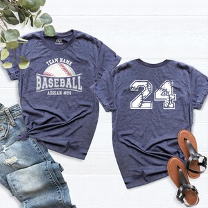 Może przedstawiać: Koszulka w kolorze melanżowego granatu z grafiką baseballową oraz napisem TEAM NAME, BASEBALL i ADRIAN #24 z przodu, a także numerem 24 z wzorem szwów piłki baseballowej z tyłu.
