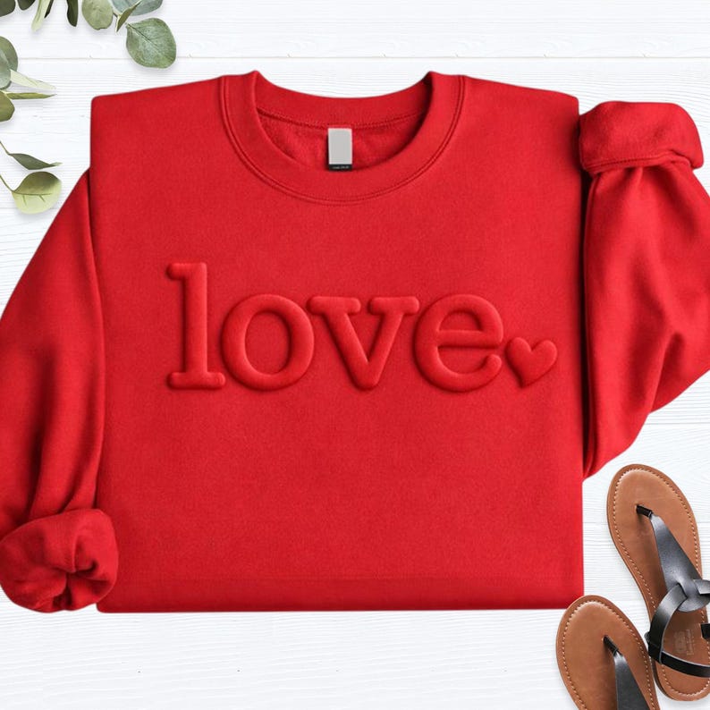 May include: A red sweatshirt with the word "love" embossed across the front. The word "love" is in a bold, sans-serif font, with a small heart replacing the "o". The sleeves are cuffed. Sandals are visible in the bottom right corner.